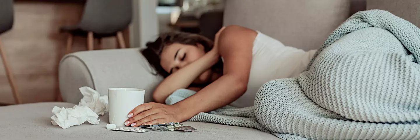 Kranke Frau liegt auf dem Sofa unter einer Decke, daneben Taschentücher, Tasse und Medikamente.