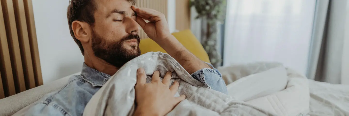 Mann mit Bart und Tattoo im Bett und hält die Hand an der Schläfe