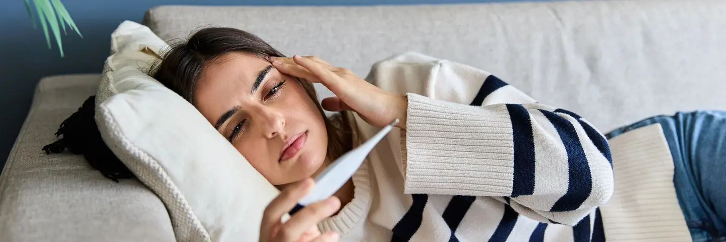 Eine Frau liegend auf dem Sofa betrachtet den Fieberthermometer
