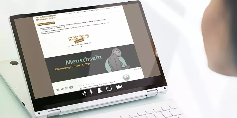 Aufsicht auf ein Laptop mit geöffneter Website des Archäologischen Museums Frankfurt, die sich eine junge Frau anschaut