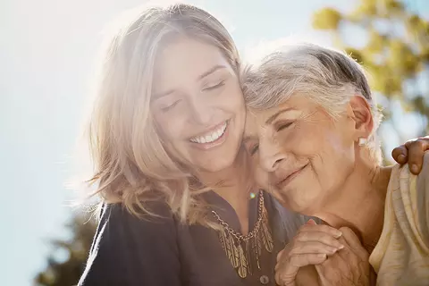 Eine jüngere Frau hält eine ältere Frau im Arm. Beide haben die Augen geschlossen und lächeln.
