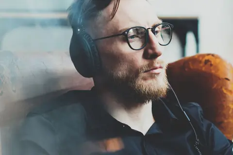 Ein junger Mann mit Bart und Brille sitzt in einem alten Ledersessel und hört mit Kopfhörer einen Podcast