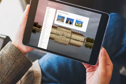 Aufsicht auf ein Tablet mit geöffneter Website des virtuellen Museums-Angebots der Bayrischen Schlösserverwaltung, dass eine junge Frau in den Händen hält