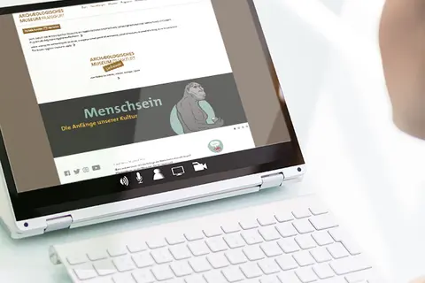 Aufsicht auf ein Laptop mit geöffneter Website des Archäologischen Museums Frankfurt, die sich eine junge Frau anschaut