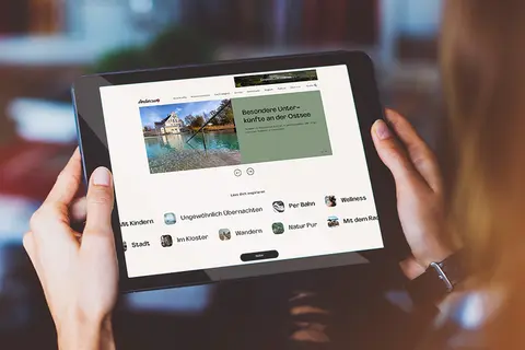 Aufsicht auf ein Tablet mit der Website wirsindanderswo.de, dass eine junge Frau in den Händen hält