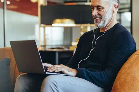 Mann sitzt auf einem Sessel und hört über Kopfhörer einen Podcast auf seinem Laptop