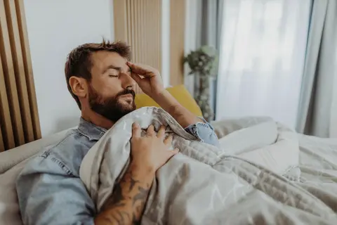 Mann mit Bart und Tattoo im Bett und hält die Hand an der Schläfe