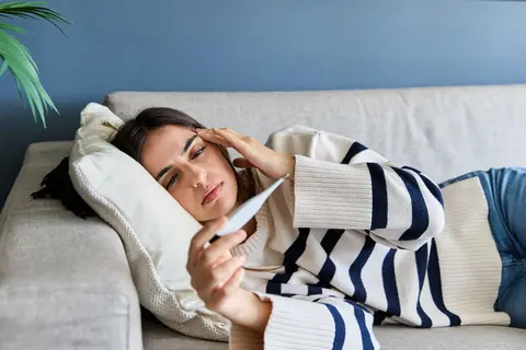 Eine Frau liegend auf dem Sofa betrachtet den Fieberthermometer