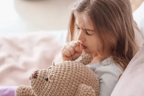 Ein Mädchen hält einen Kuschelbär in der Hand und hustet