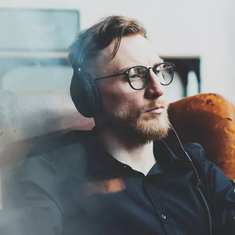 Ein junger Mann mit Bart und Brille sitzt in einem alten Ledersessel und hört mit Kopfhörer einen Podcast