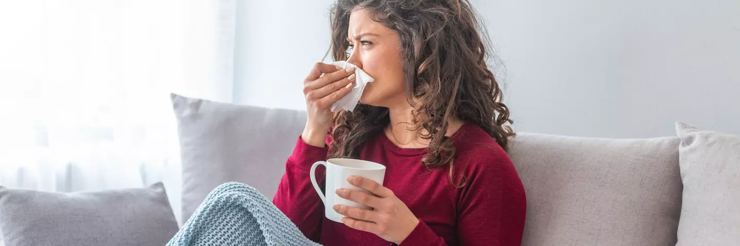 Frau sitzt auf dem Sofa, hält eine Tasse und schnäuzt sich mit einem Taschentuch.