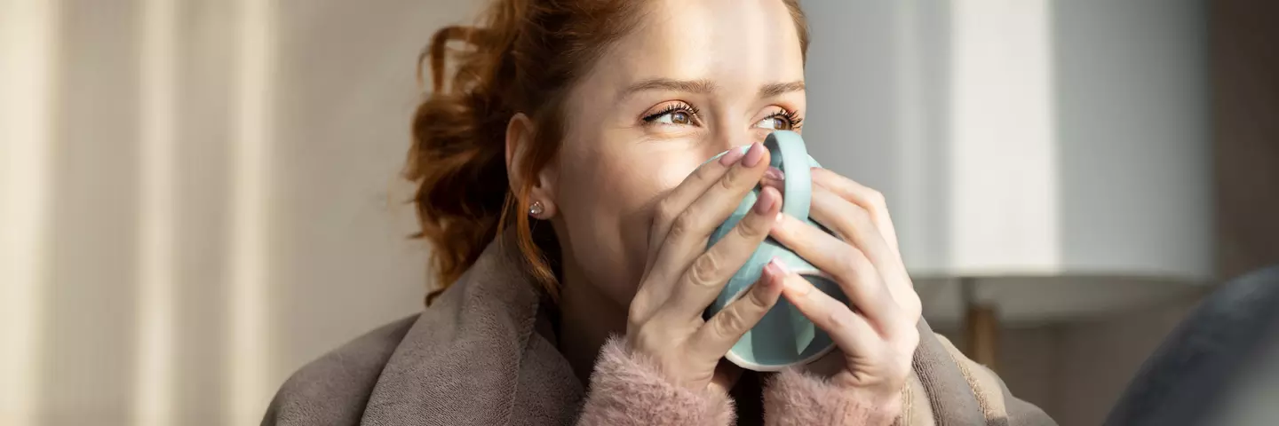 Frau mit hochgebundenen Haaren trinkt aus einer großen Tasse und blickt lächelnd aus dem Fenster.