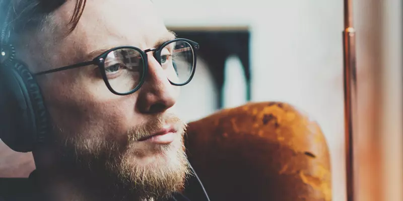 Ein junger Mann mit Bart und Brille sitzt in einem alten Ledersessel und hört mit Kopfhörer einen Podcast