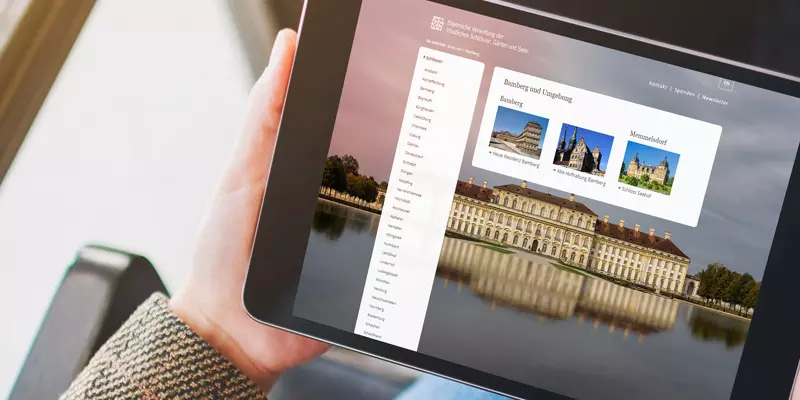 Aufsicht auf ein Tablet mit geöffneter Website des virtuellen Museums-Angebots der Bayrischen Schlösserverwaltung, dass eine junge Frau in den Händen hält