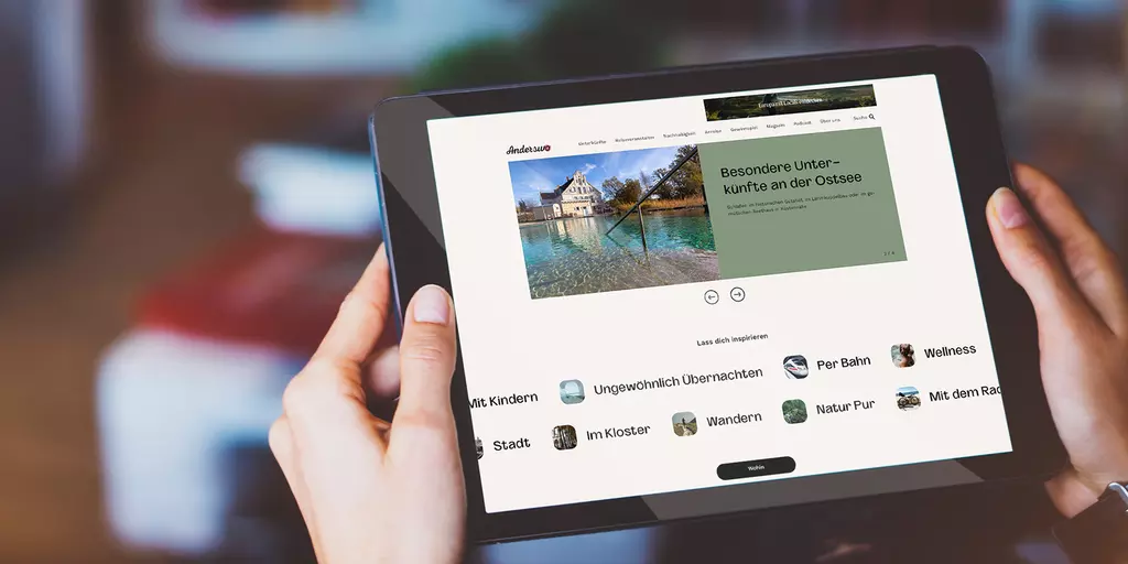Aufsicht auf ein Tablet mit der Website wirsindanderswo.de, dass eine junge Frau in den Händen hält