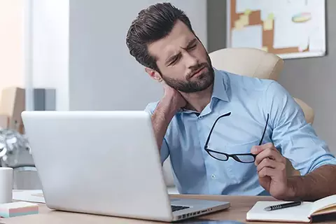 Junger Mann sitzt am Laptop und greift sich wegen Nackschmerzen ans Genick