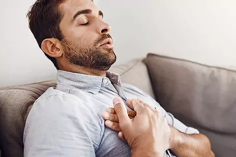 Mann sitzt auf dem Sofa und hält sich wegen Sodbrennen die Brust