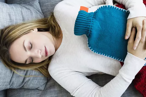 Frau liegt mit Bauchschmerzen wegen Verdauungsstörungen mit einer Wärmflasche auf dem Sofa