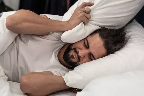 Mann mit Schlafstörungen liegt im Bett und hält sich Kissen auf die Ohren