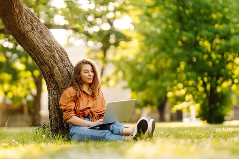 Eine junge Frau sitzt auf einer Wiese an einen Baum gelehnt und schaut auf ihren Laptop, den sie auf dem Schoß hat