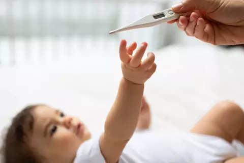 Hand hält ein digitales Thermometer über einem liegenden Baby, das nach dem Thermometer greift.