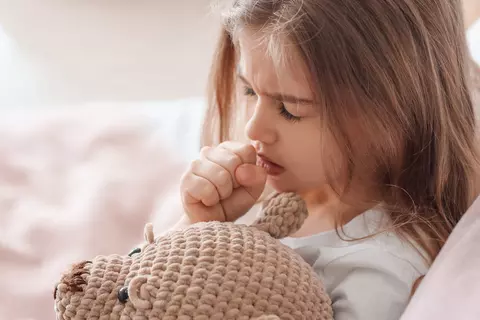 Kleines Mädchen liegt im Bett, hält einen gehäkelten Teddybären und hustet in die Faust.