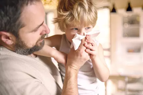 Vater hält ein kleines Kind im Arm, das sich mit einem Taschentuch die Nase putzt.
