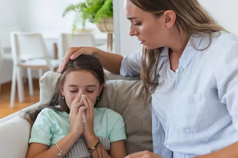 Mutter sitzt neben ihrer kranken Tochter auf dem Sofa und legt beruhigend die Hand auf ihren Kopf.