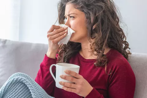 Frau sitzt auf dem Sofa, hält eine Tasse und schnäuzt sich mit einem Taschentuch.
