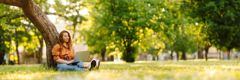 Eine junge Frau sitzt auf einer Wiese an einen Baum gelehnt und schaut auf ihren Laptop. den sie auf dem Schoß hat