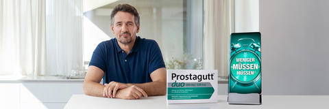 Mann sitzt am Tisch neben einer Prostagutt Packung und einem überdimensionierten Mobiltelefon mit dem Slogan: Weniger müssen müssen