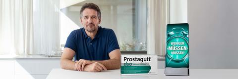 Mann sitzt am Tisch neben einer Prostagutt Packung und einem überdimensionierten Mobiltelefon mit dem Slogan: Weniger müssen müssen