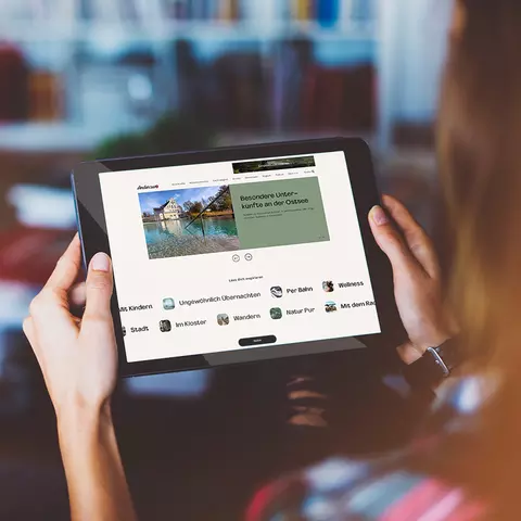 Aufsicht auf ein Tablet mit der Website wirsindanderswo.de, dass eine junge Frau in den Händen hält