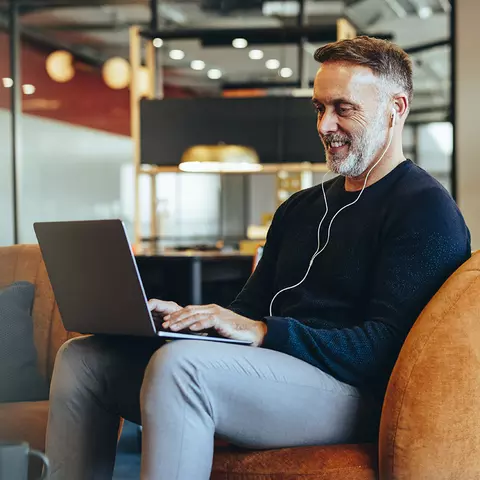 Mann sitzt auf einem Sessel und hört über Kopfhörer einen Podcast auf seinem Laptop