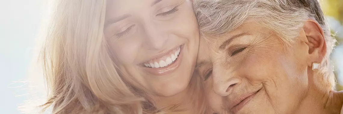 Eine jüngere Frau hält eine ältere Frau im Arm. Beide haben die Augen geschlossen und lächeln.