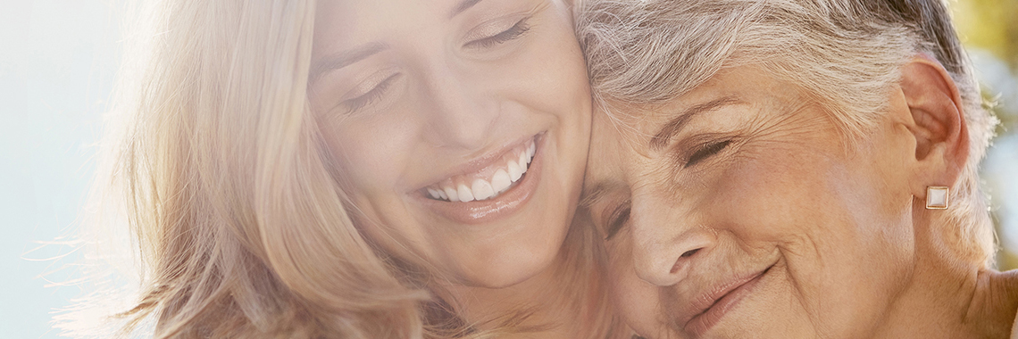 Eine jüngere Frau hält eine ältere Frau im Arm. Beide haben die Augen geschlossen und lächeln.